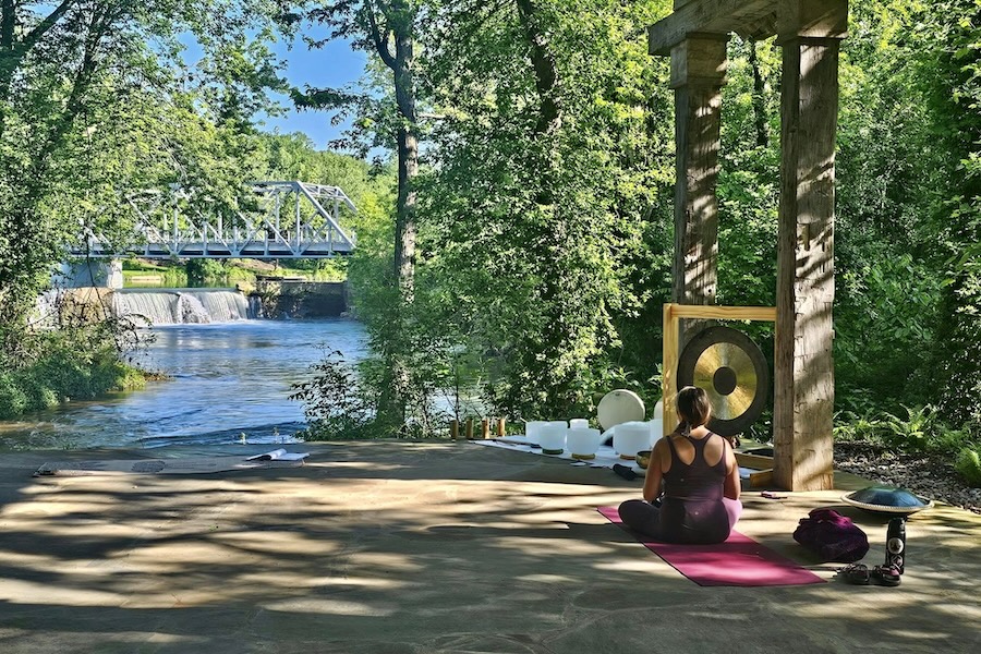 A woman sits on a yoga mat next to a gong overlooking a river with a bridge going over it