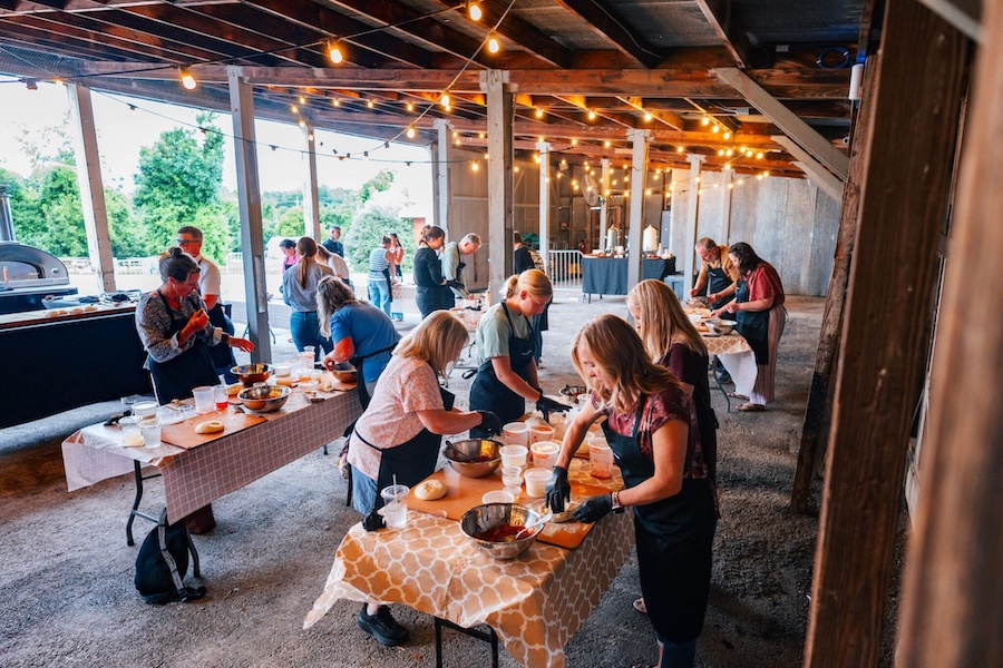 Group learns to make pizza in a rustic outdoor setting under a covered pavilion