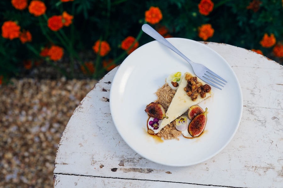 A slice of cheese cake topped with candied nuts and surrounded by figs and flowers on a white wooden table outdoors