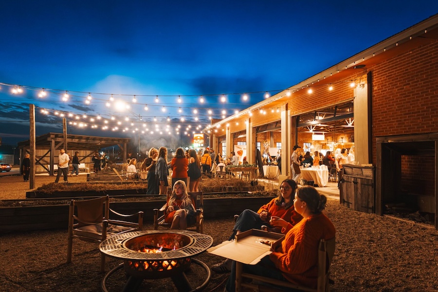 People gather around fire pits and rtables at nightfall under festooned Edison bulb lights next to a brick building with open barn doors to allow the cool night air in