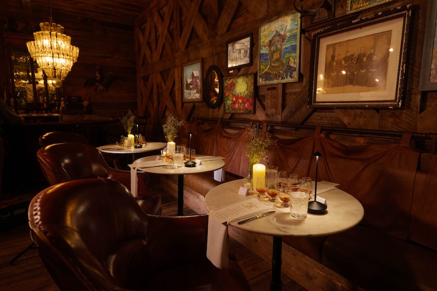Dimly lit dining room with round tables agains a wall length booth bench with chandeliers and candles shining on folk art paintings on wooden walls