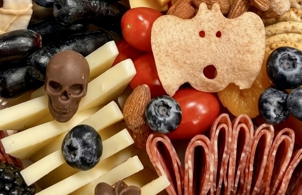 Charcuterie food arranged on a wooden board with berries, tomatoes, meats, and Halloween themed snacks with cheese ranged in the shape of a spine or rib cage
